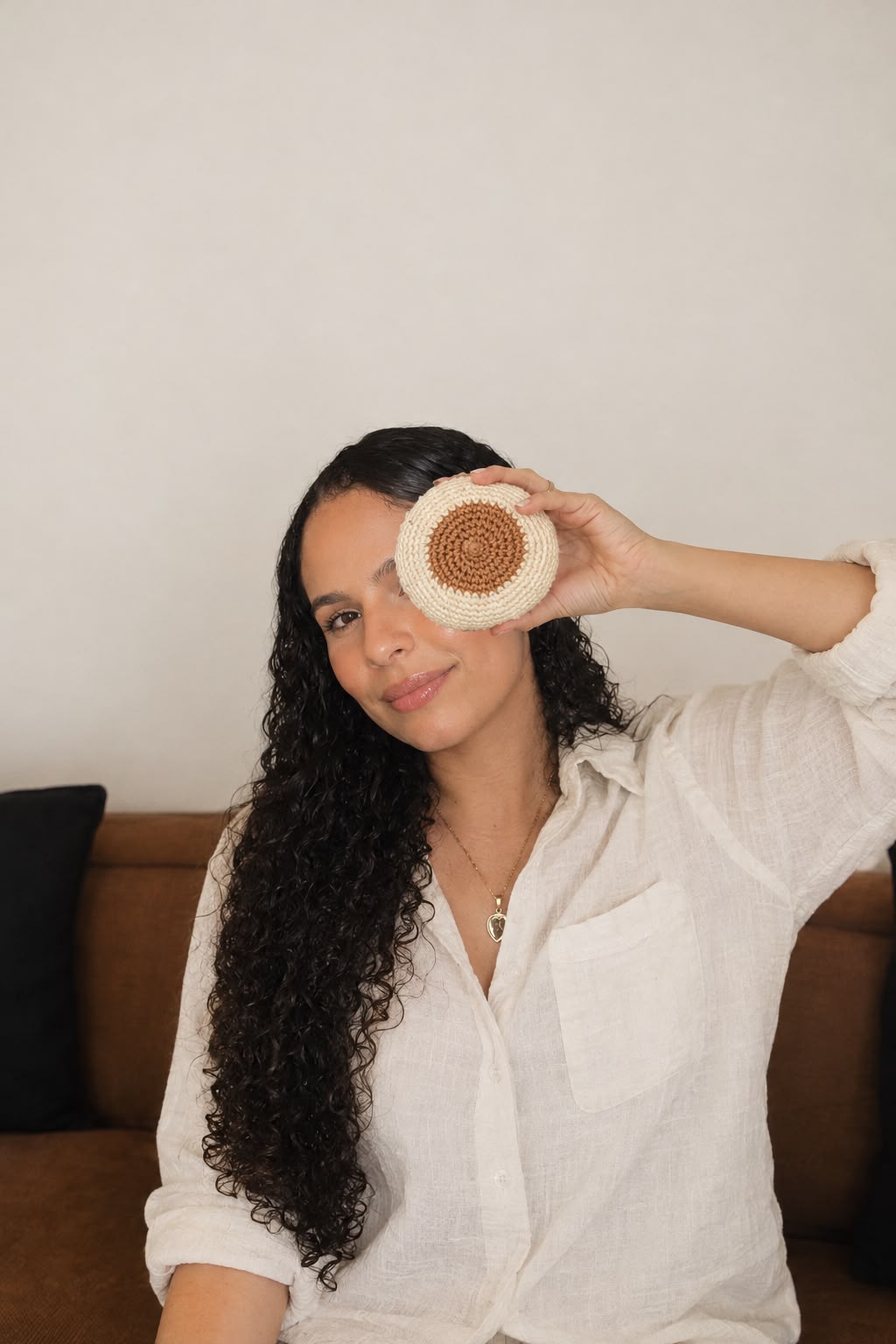 Jakeline Barbosa sorri sentada em um sofá, com uma das mãos erguida segurando uma peça circular de crochê diante de um dos olhos.