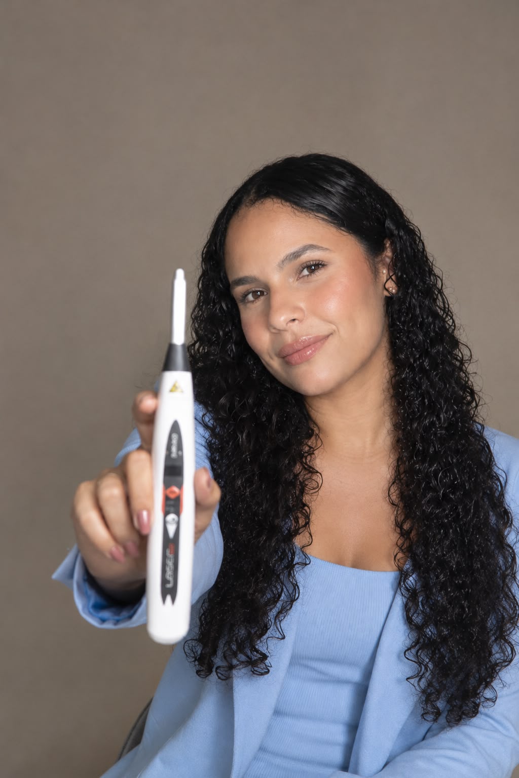 Enfermeira Jakeline Barbosa, de blazer azul-claro, sorri e segura à frente um aparelho portátil de laserterapia.
